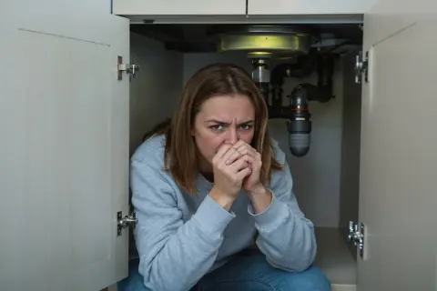 Femme inquiète sous un évier, face à un **tout à l'égout bouché que faire** ? Les tuyaux sont visibles.