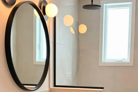 Un élégant miroir au-dessus du lavabo reflète des lumières douces. La salle de bain moderne comprend une douche vitrée et une fenêtre lumineuse.