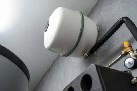 Vue rapprochée du **vase d'expansion chauffage**, élément blanc fixé au mur, avec tuyauterie noire isolée et raccords en laiton.