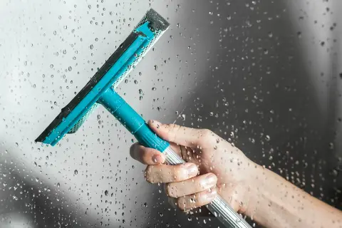 Main tenant une raclette bleue pour le nettoyage vitre salle de bain, laissant des traces d'eau sur la paroi.