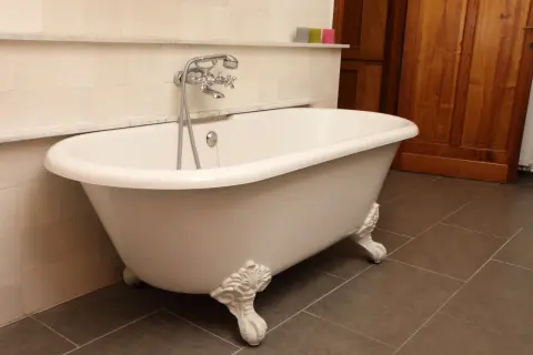 Baignoire îlot blanche sur pieds sculptés, évoquant le poids d'une baignoire en fonte. Robinet argenté, carrelage mural clair et sol sombre.