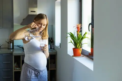 Une femme enceinte verse de l'eau du robinet dans un verre. Elle est dans sa cuisine, près d'une fenêtre avec une plante.
