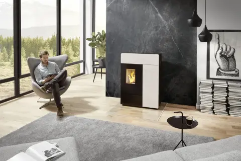 Un homme lit dans un fauteuil près d'un poêle moderne. Le **carrelage derrière le poêle** en marbre noir crée un contraste saisissant.