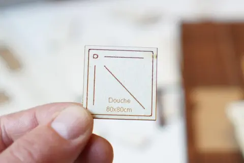 Main tenue par un pouce, une petite carte en bois gravée indique "Douche 80x80cm", une dimension pour une future douche.