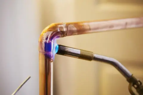 Un chalumeau chauffe un raccord coudé en cuivre pour le souder. La flamme bleue et violette illumine le métal.