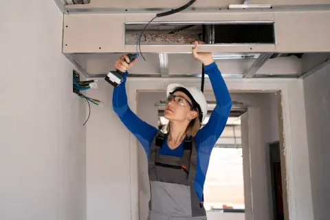 Une femme en casque et lunettes, avec une perceuse, installe un faux plafond pour un système de climatisation split gainable.