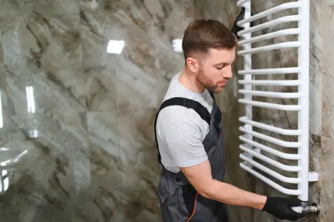Un plombier en salopette ajuste la vanne d'un sèche serviette blanc, prêt pour son installation.