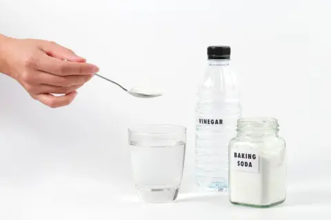 Une main tient une cuillère de bicarbonate de soude pour déboucher une canalisation. Vinaigre et eau sont prêts.