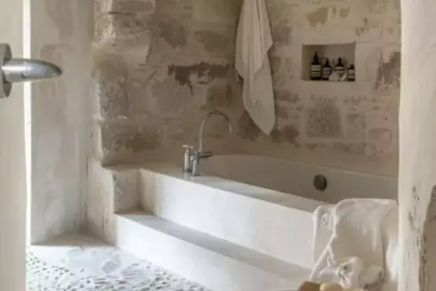 Une **salle de bain méditerranéenne** avec baignoire en pierre brute, robinetterie moderne et serviette blanche suspendue.