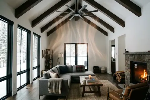 Salon cosy avec cheminée en pierre, canapé gris, grandes fenêtres donnant sur la neige. Le **chauffage au plafond** discret assure une chaleur douce.